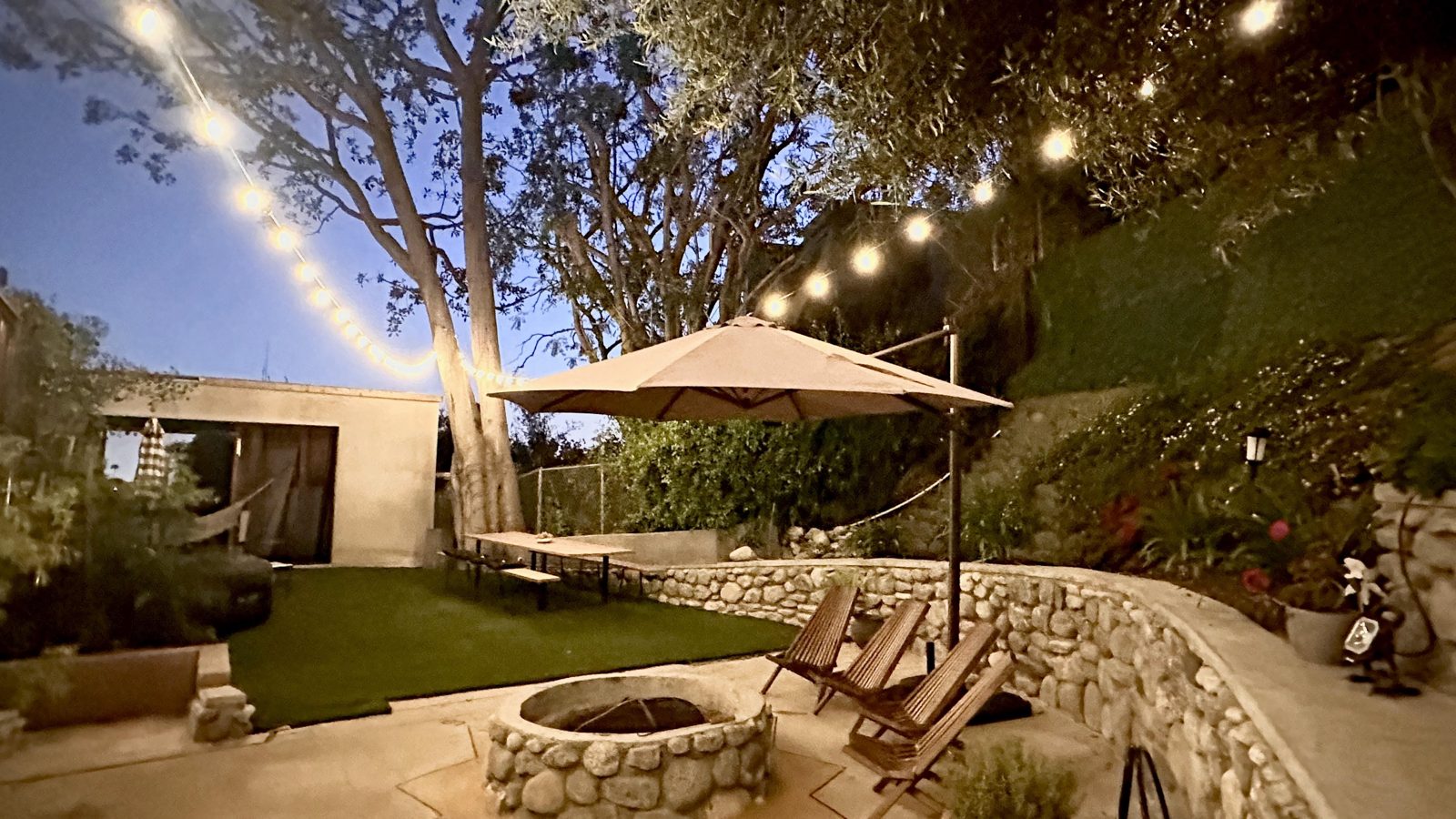 The stone fire pit at dusk, adirondack chairs and string lights in the shared backyard